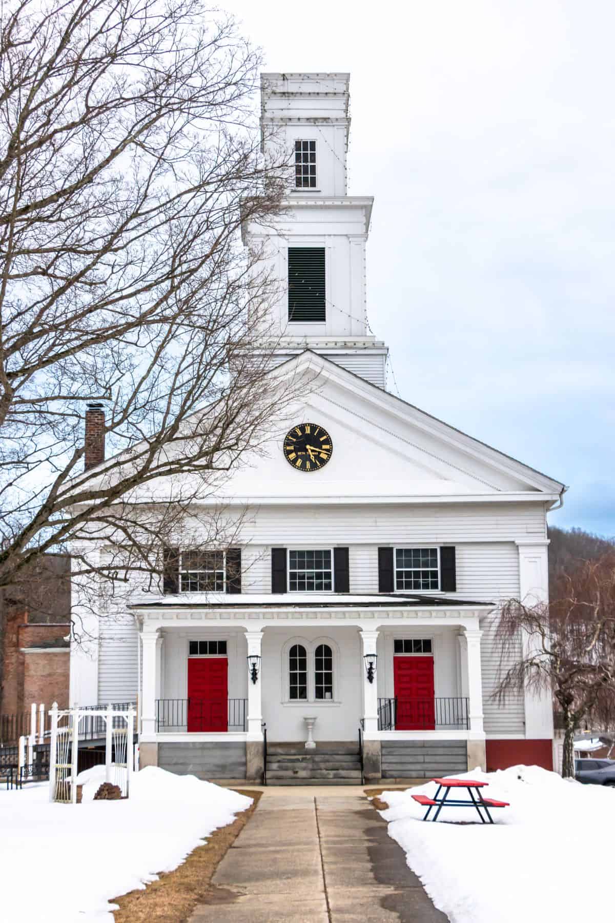 A white church with a central clock tower, black shutters, and three red doors, surrounded by snow with bare trees and a picnic table in front.