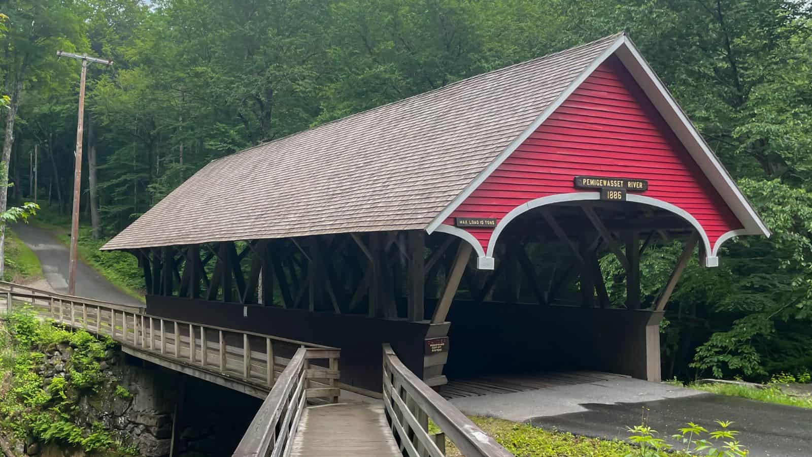 A red wooden covered bridge labeled "Pemigewasset 1886" spans a small road in a forested area, with a wooden walkway leading up to it.