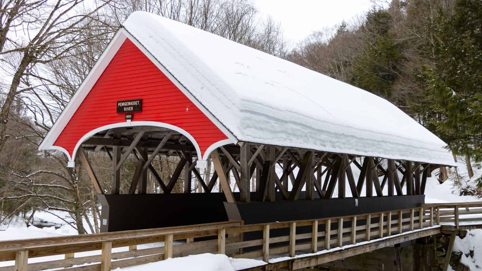 A red covered bridge with a snow-covered roof spans over a creek, surrounded by trees in a winter landscape.