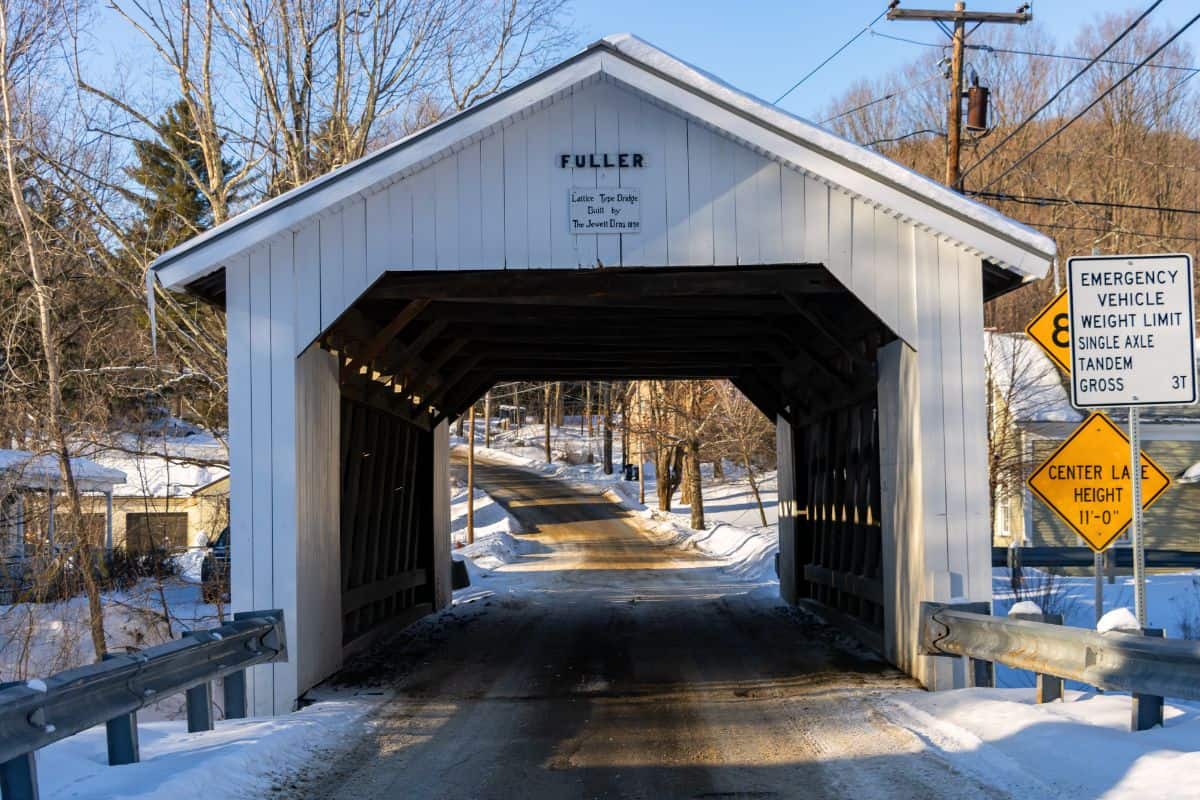 A white wooden covered bridge named Fuller spans a snow-lined road, with road signs indicating vehicle weight and height limits at the entrance.