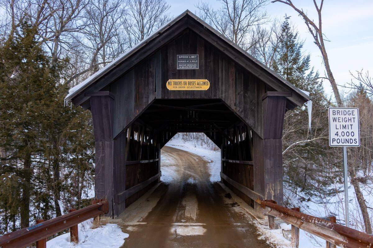 A wooden covered bridge with snow on the ground, displaying signs for speed, weight limit, and truck restrictions; leafless trees surround the scene.