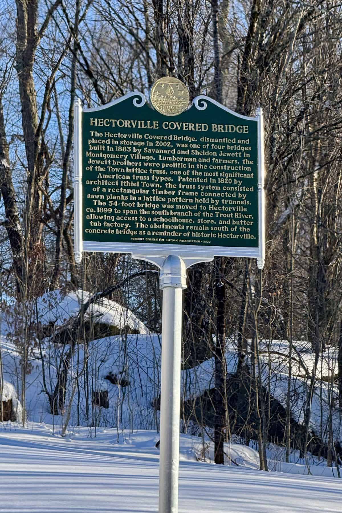Green and white historical marker titled "Hectorville Covered Bridge" stands in a snowy, wooded area, detailing the bridge's history and relocation.