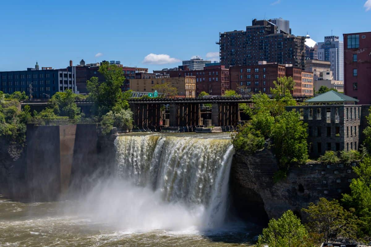A waterfall flows over a rocky ledge in an urban area, with mist rising and buildings in the background under a clear blue sky.
