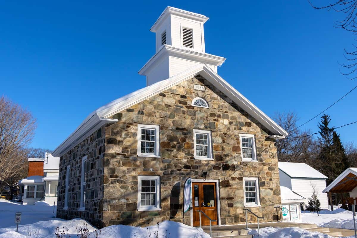 A historic stone building with a white bell tower stands amid snow under a clear blue sky; date 1844 is visible above the entrance.
