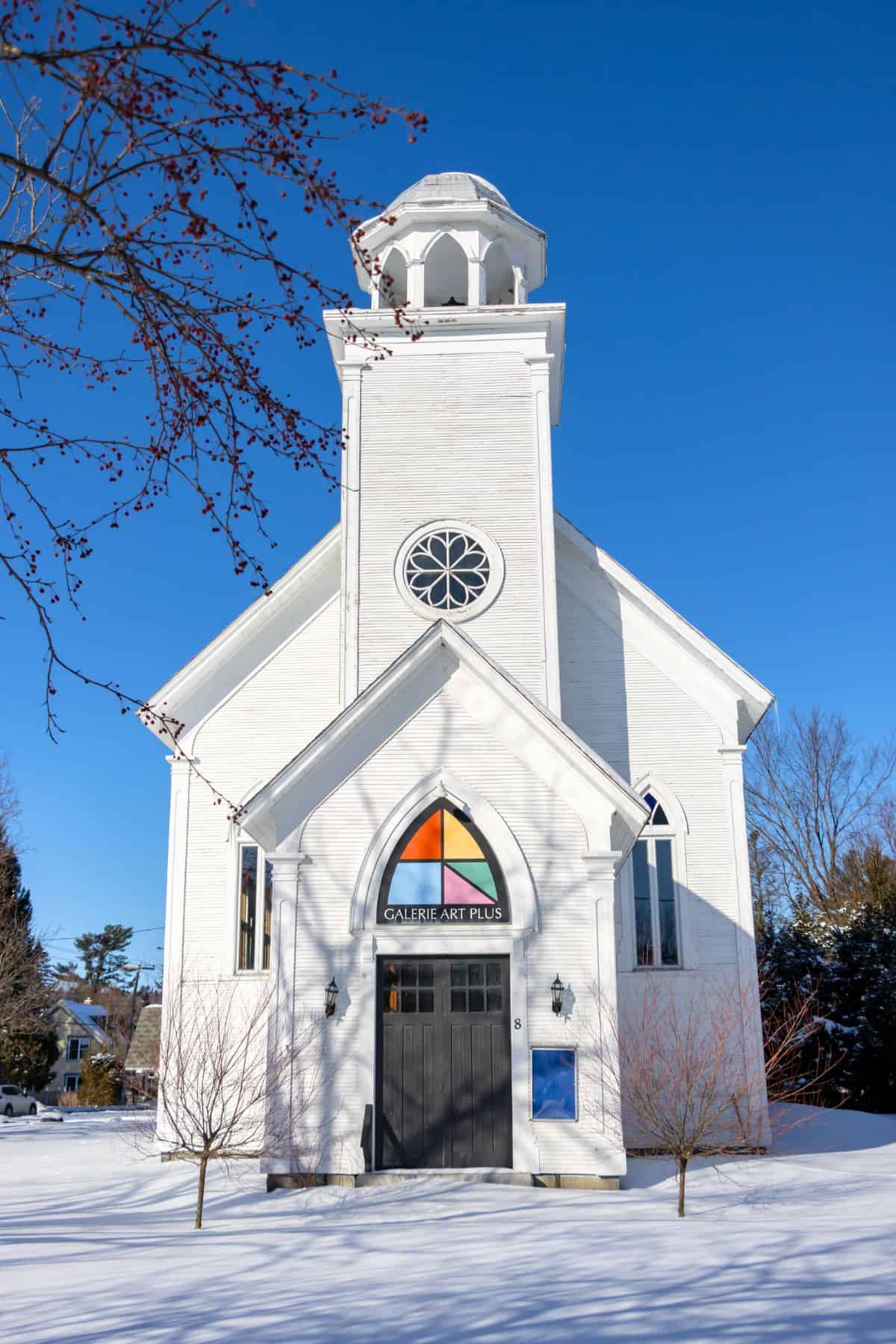 White church building with a steeple, now serving as an art gallery. Snow covers the ground, and the sky is clear and blue.