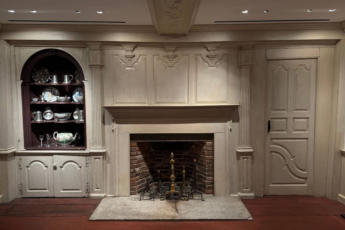 A traditional room with a central brick fireplace, ornate wooden mantel, closed panel door on the right, and a cabinet with shelves displaying dishes on the left.