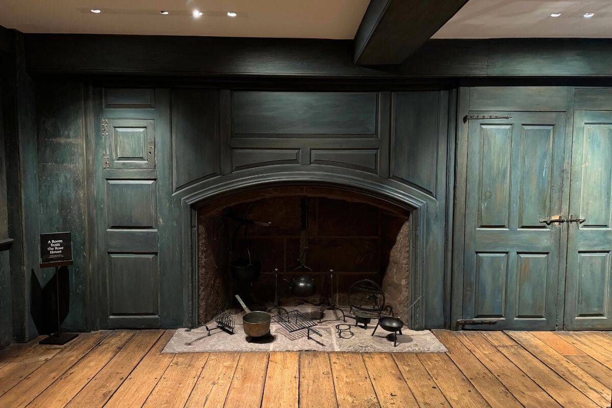 A large, old-fashioned fireplace with green wooden paneling, various antique fireplace tools, and a wooden floor in a dimly lit room.
