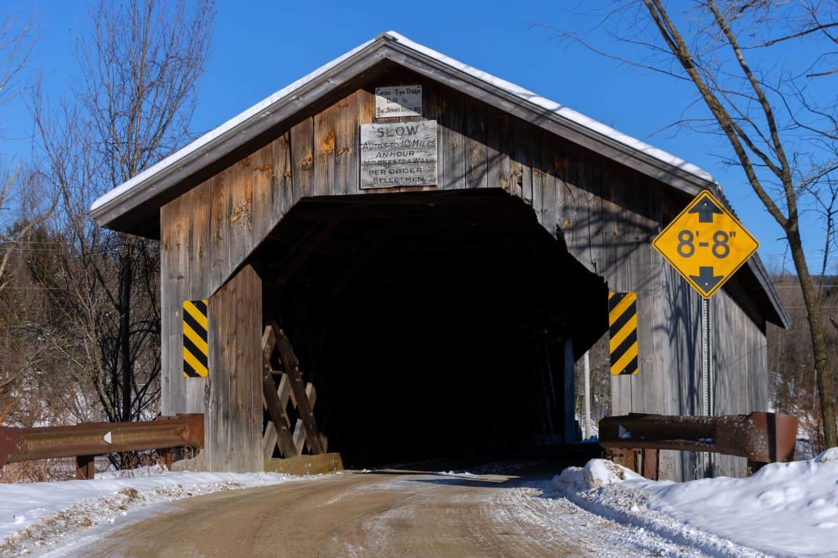 A wooden covered bridge with a snow-covered dirt road passing through, height restriction sign on the right, and caution signs on the bridge.