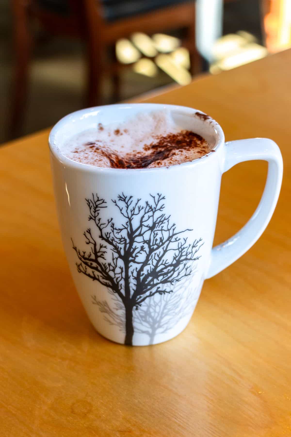 A white mug with a black tree design holds a frothy drink topped with cocoa powder, sitting on a wooden table.