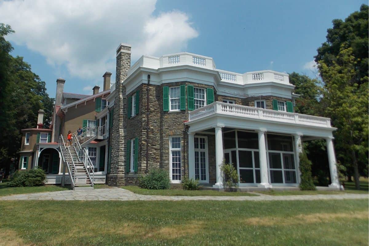 Large historic house with stone walls, green shutters, multiple chimneys, and a white columned porch. Two people are on the exterior staircase. Grass and trees surround the building.
