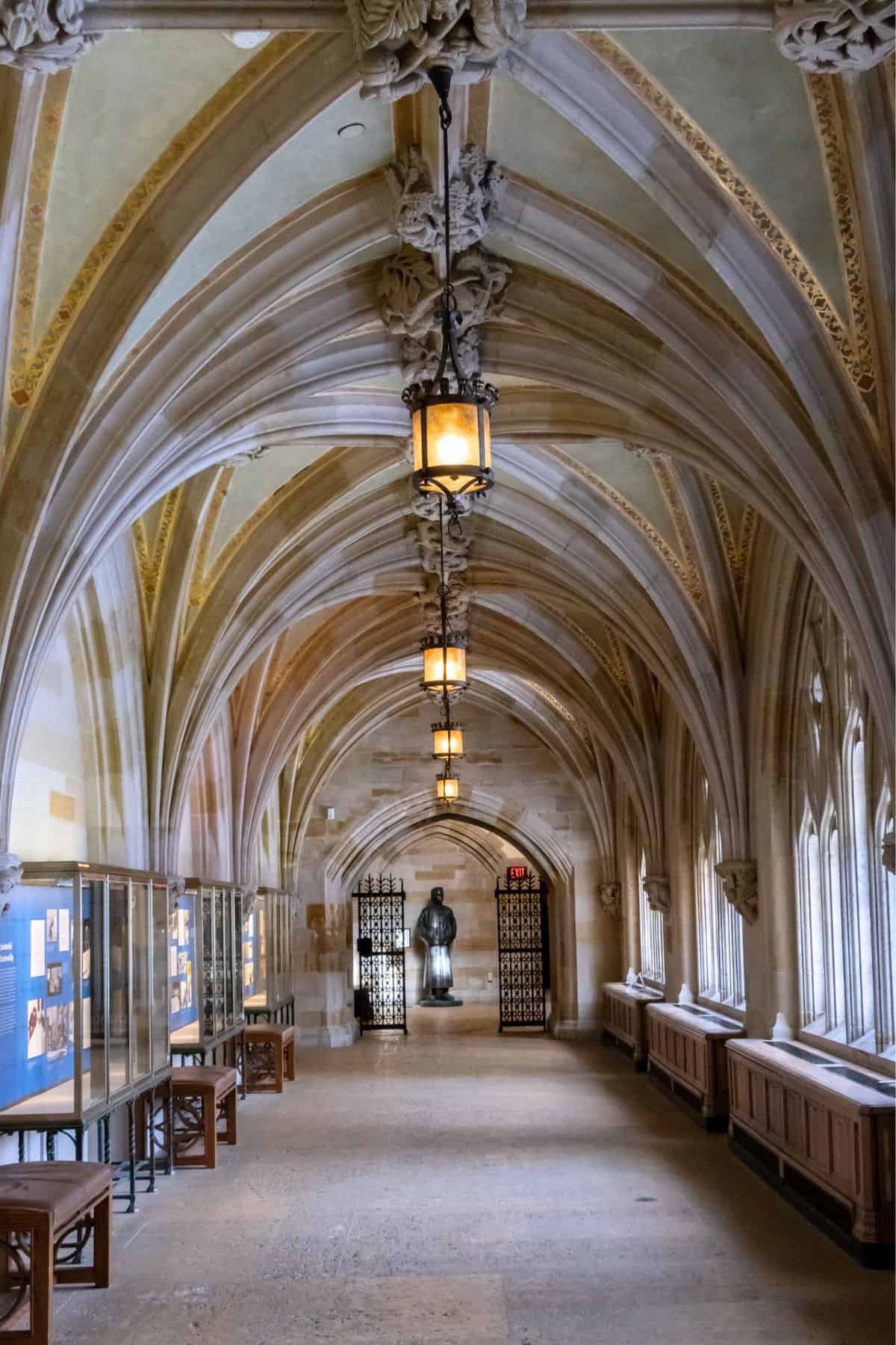 A stone hallway with vaulted gothic arches, hanging lanterns, display cases on the left, arched windows on the right, and a statue in front of a metal gate at the far end.