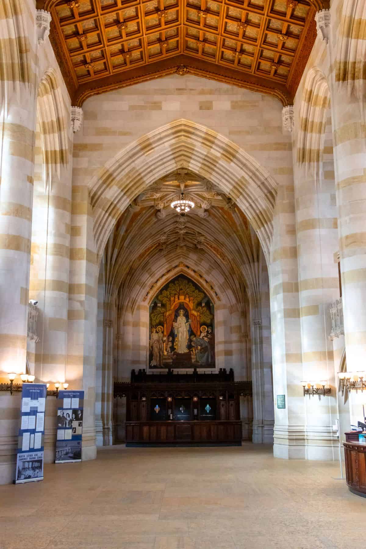 A grand hall with high vaulted ceilings, striped stone walls, wooden ceiling detail, arched entry, and a mural above a dark wooden structure at the far end.