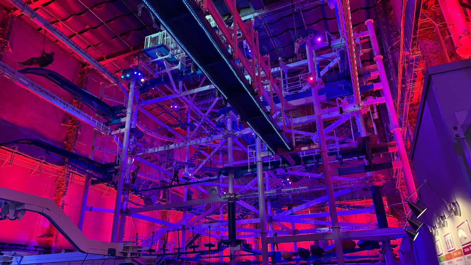 Intricate indoor ropes course and obstacle structure lit with red, purple, and blue lights, featuring beams, cables, platforms, and elevated walking tracks.