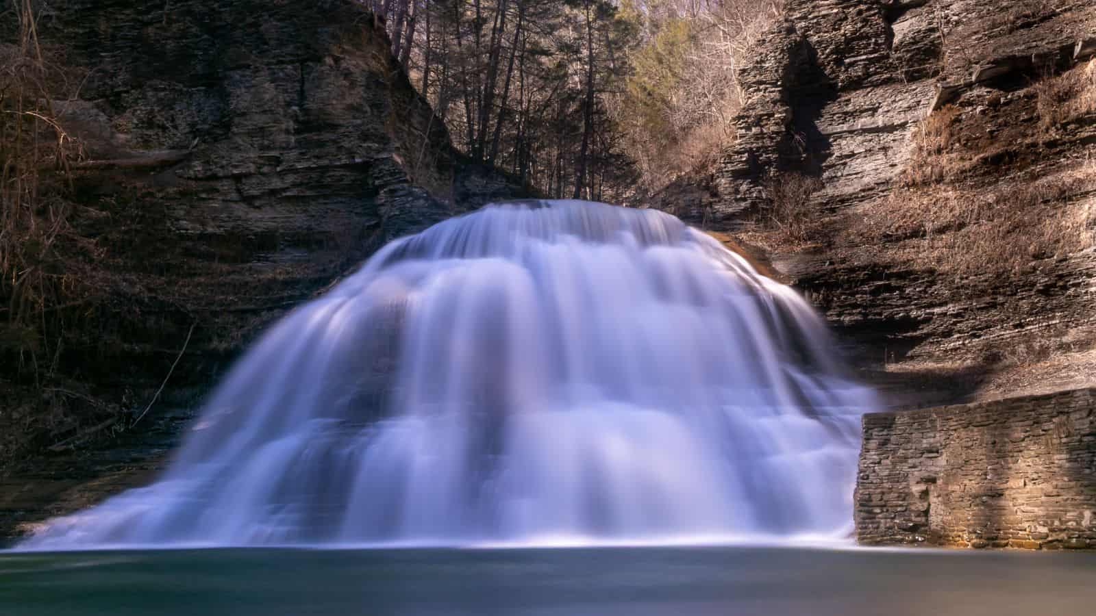 A wide waterfall cascades over rocky ledges into a calm pool, surrounded by steep, layered rock walls and sparse trees.