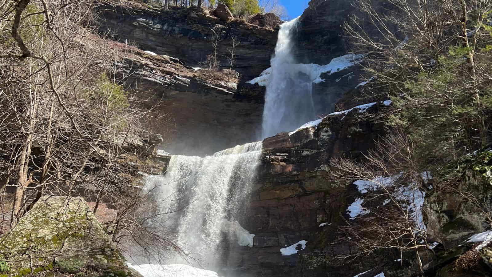 A two-tiered waterfall flows over rocky cliffs, surrounded by leafless trees and patches of snow on a sunny day.