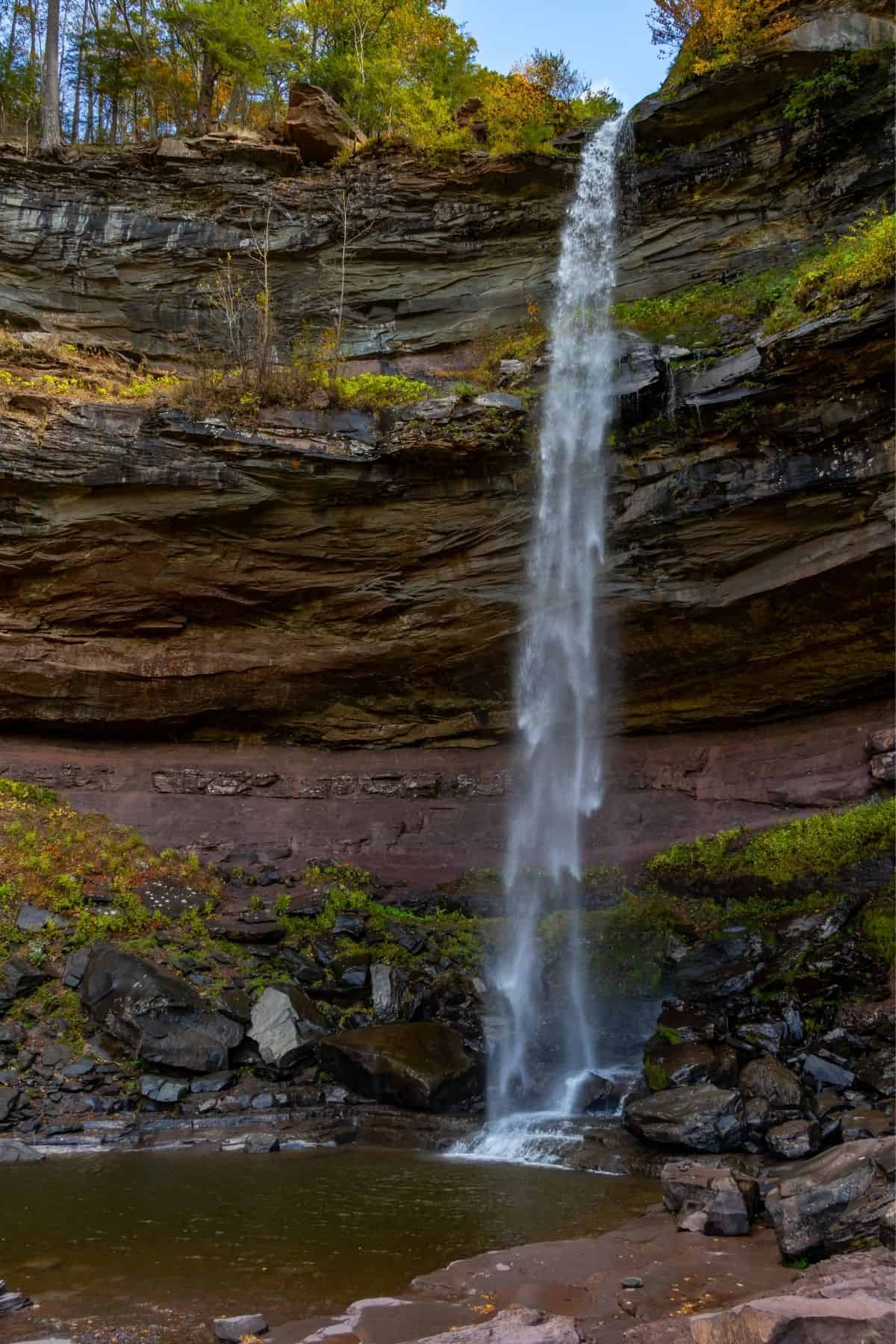 A tall, narrow waterfall cascades from a rocky cliff into a small pool below, surrounded by rocks and greenery.