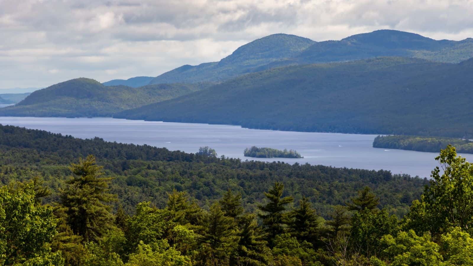 View of a large lake surrounded by dense green forest and rolling mountains under a cloudy sky.