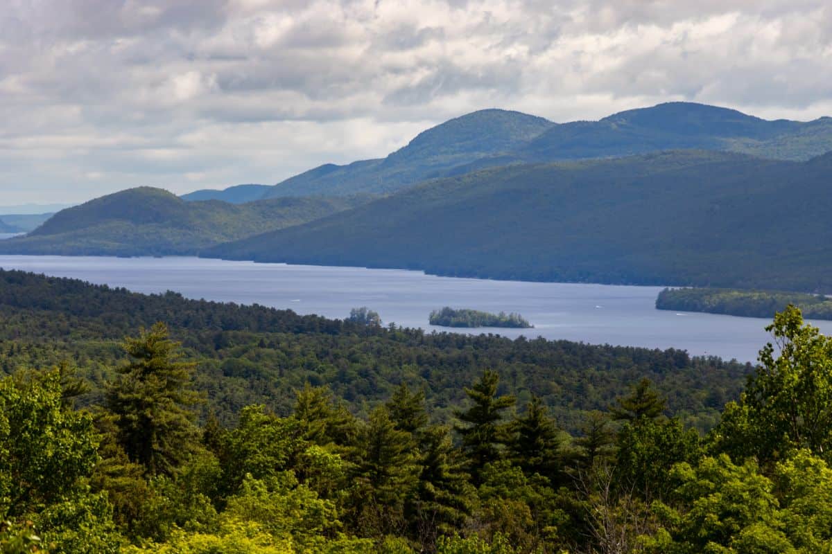 A large lake bordered by dense green forest and rolling mountains under a partly cloudy sky.