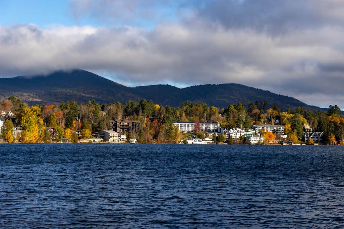 A lake with calm water in the foreground, trees with autumn foliage, buildings along the shore, and forested mountains under a cloudy sky in the background.