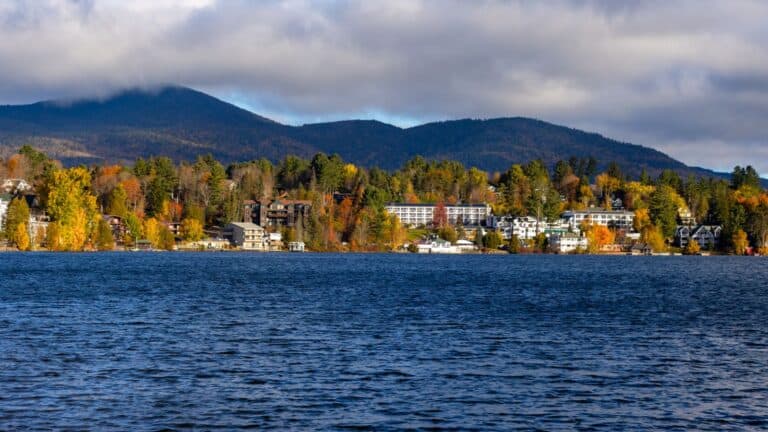A large lake with gentle waves is in the foreground; a tree-lined shore with buildings and autumn foliage sits in front of rolling mountains under a cloudy sky.