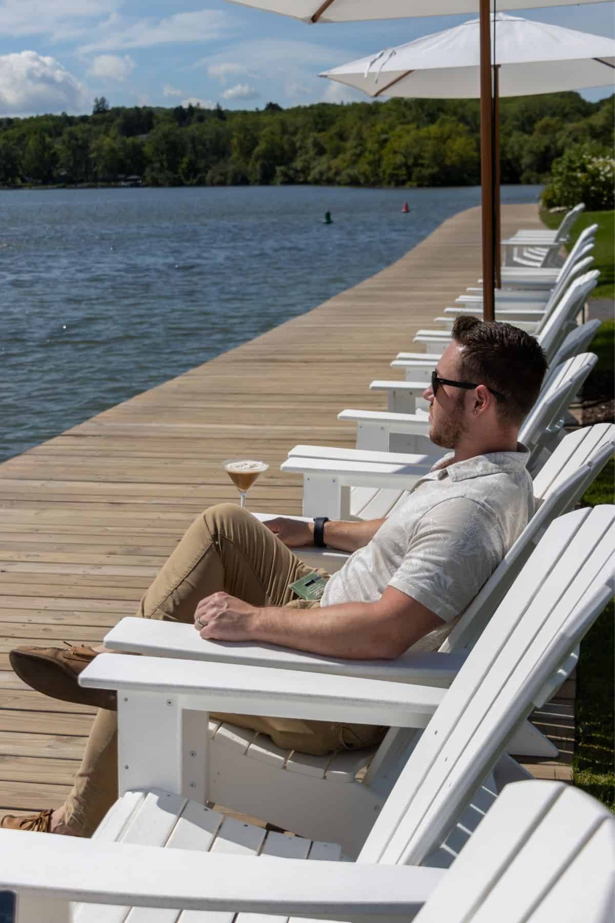 Man in sunglasses sits on a white chair by a lakeside dock, holding a cocktail. Umbrellas and more empty chairs line the wooden dock on a sunny day.