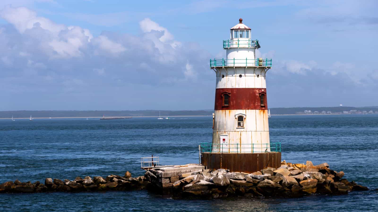 A cylindrical lighthouse with a red and white exterior sits on a rocky breakwater surrounded by blue ocean water under a partly cloudy sky.