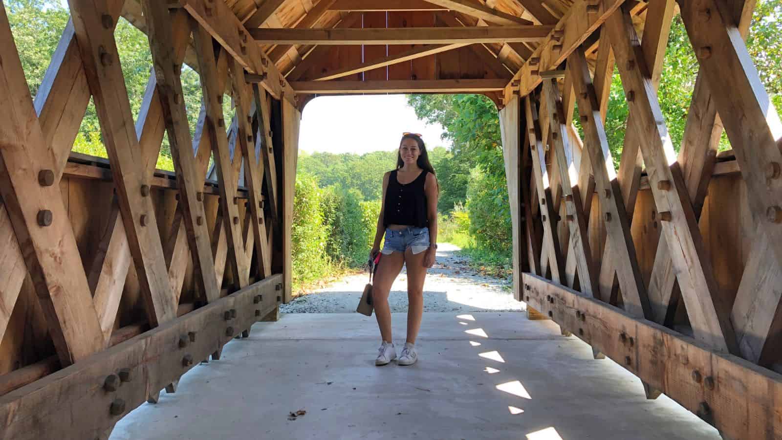 A woman stands in the middle of a covered wooden bridge, surrounded by greenery and lit by natural sunlight.