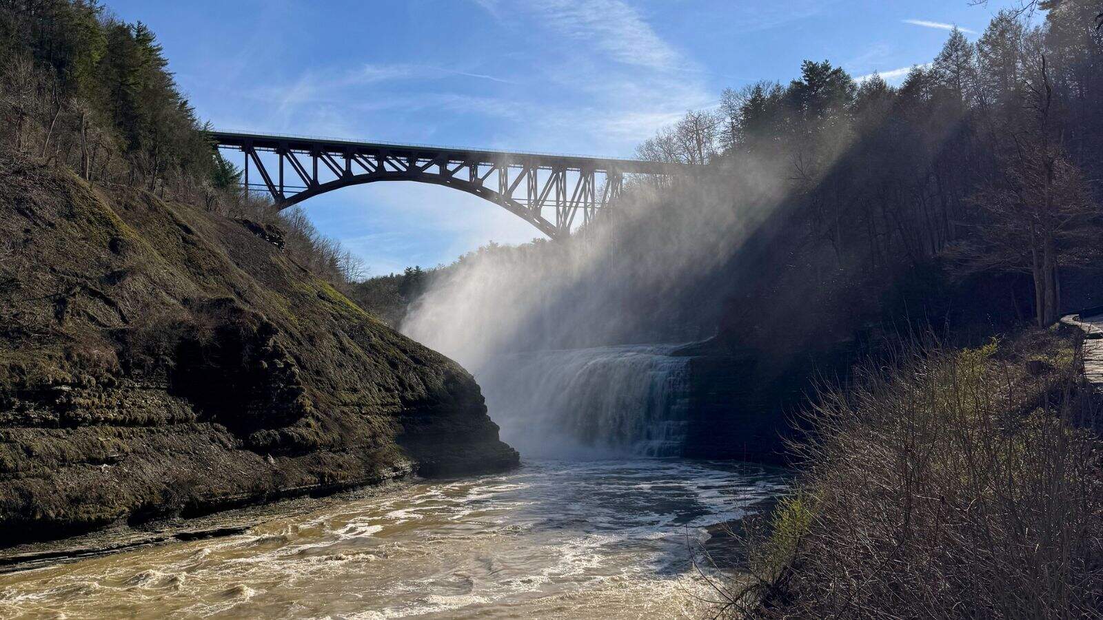 A steel bridge spans over a waterfall with mist rising, surrounded by rocky cliffs and trees under a blue sky.