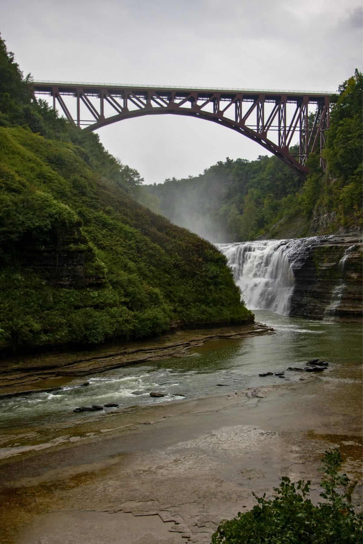 A large iron bridge spans above a waterfall surrounded by green hills, with a river flowing in the foreground under a cloudy sky.