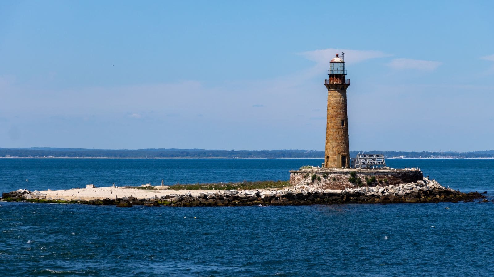 A tall lighthouse stands on a rocky island surrounded by blue water, with a small building nearby and distant land visible on the horizon.
