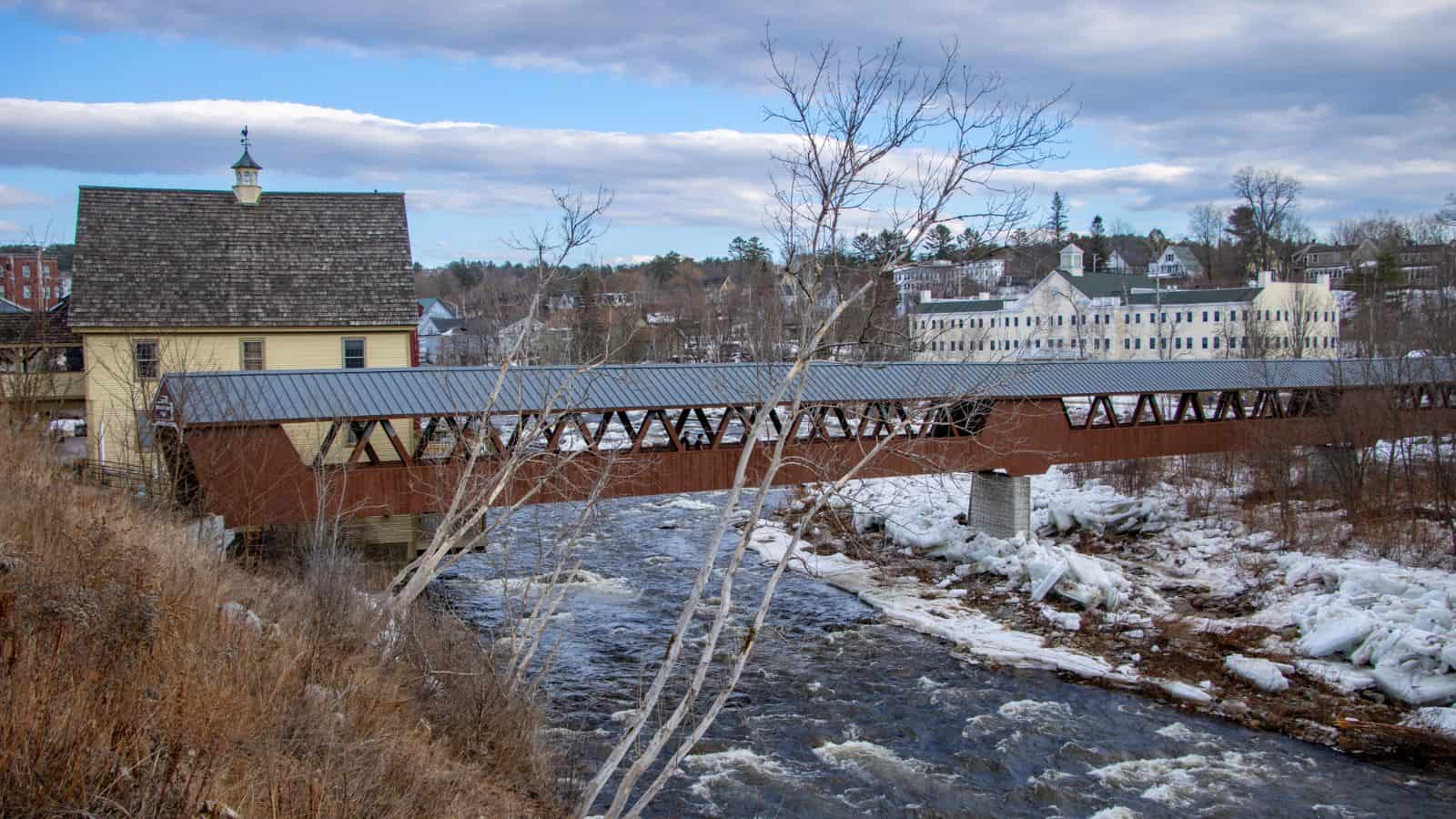 A covered wooden bridge spans a partially frozen river in a small town, with historic buildings visible in the background under a partly cloudy sky.