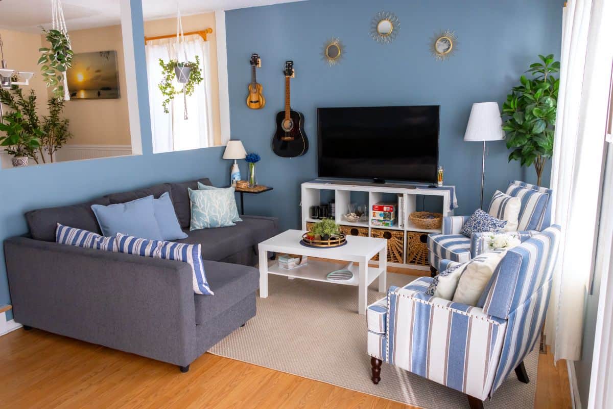 A cozy living room with blue walls, a gray sofa, striped armchairs, a TV, guitars on the wall, plants, and a white coffee table on a beige rug.