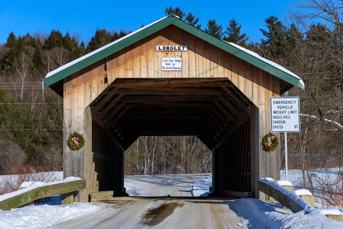 A wooden covered bridge labeled "Longley" crosses a snowy road; wreaths hang on each side, and a sign states an 8-ton emergency vehicle weight limit.