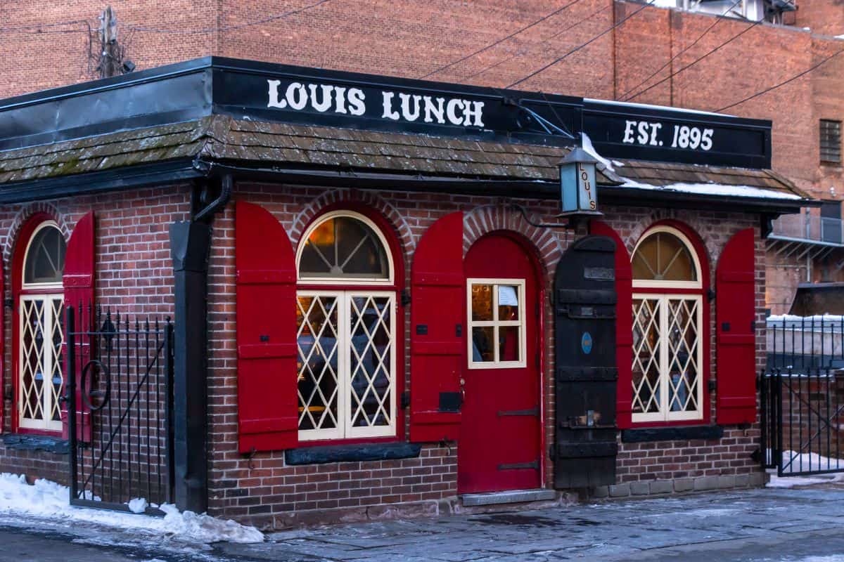 A small brick building with red doors and shutters, diamond-pane windows, and a sign reading "Louis Lunch Est. 1895" on the roof. Snow is visible on the ground.
