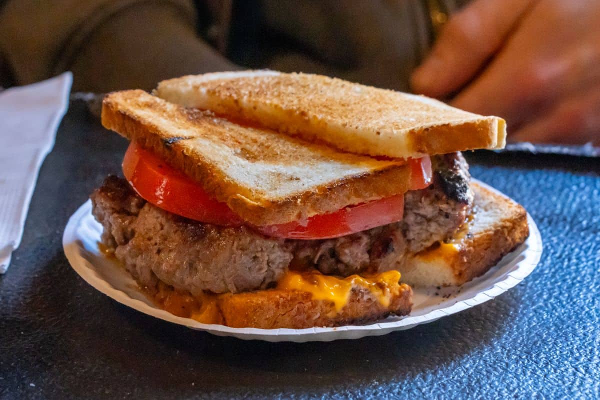 A cheeseburger with tomato slices is served between two pieces of toasted sandwich bread on a white paper plate.