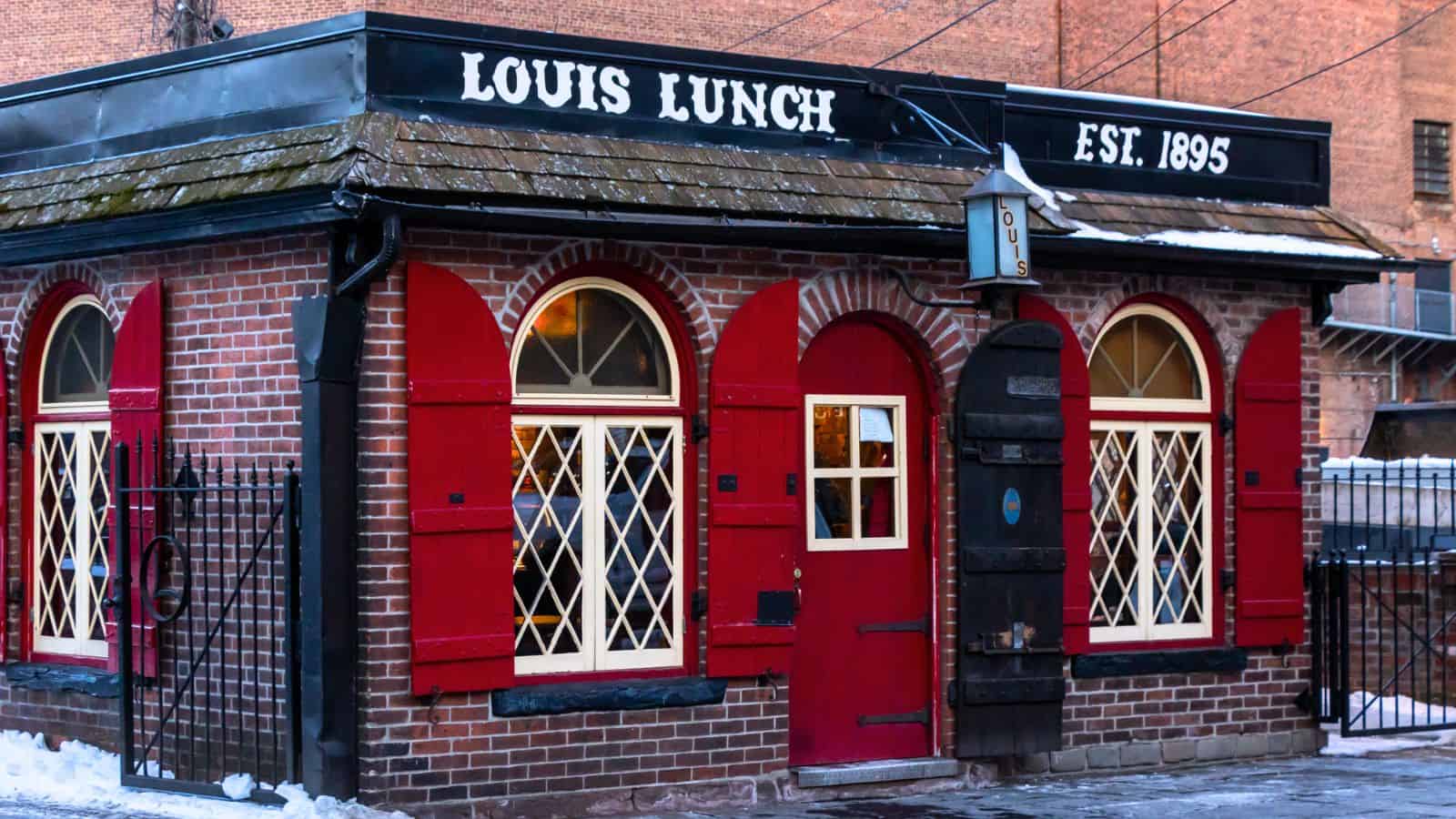 A small brick building with red shutters and a sign reading "Louis Lunch Est. 1895" above the door, surrounded by snow.