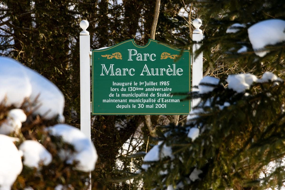 Green sign for Parc Marc Aurèle, inaugurated in July 1985, marking the 130th anniversary of Stukely, now part of Eastman since May 30, 2001; surrounded by snow-covered trees.