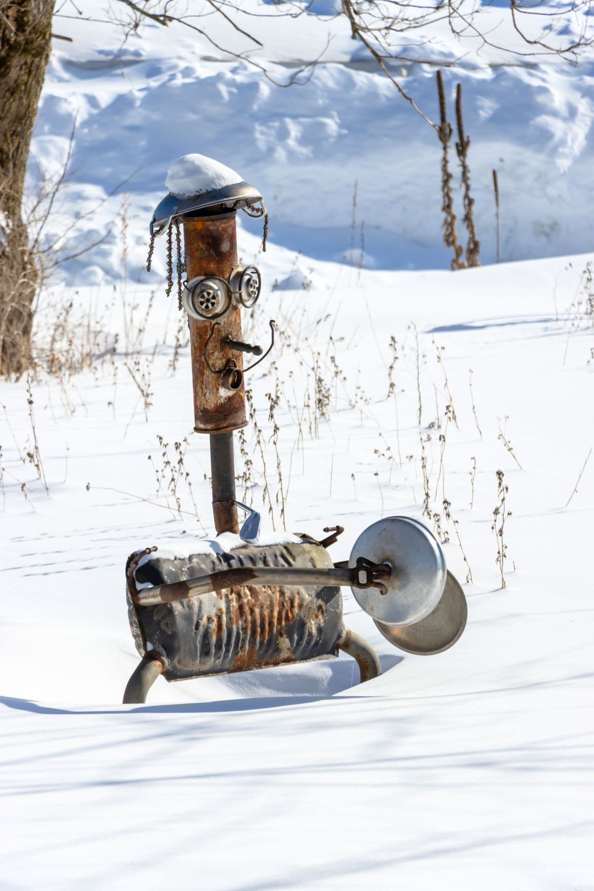 A metal sculpture resembling a humanoid figure stands in a snowy field, partially covered in snow, with various found objects and parts assembled as its features.