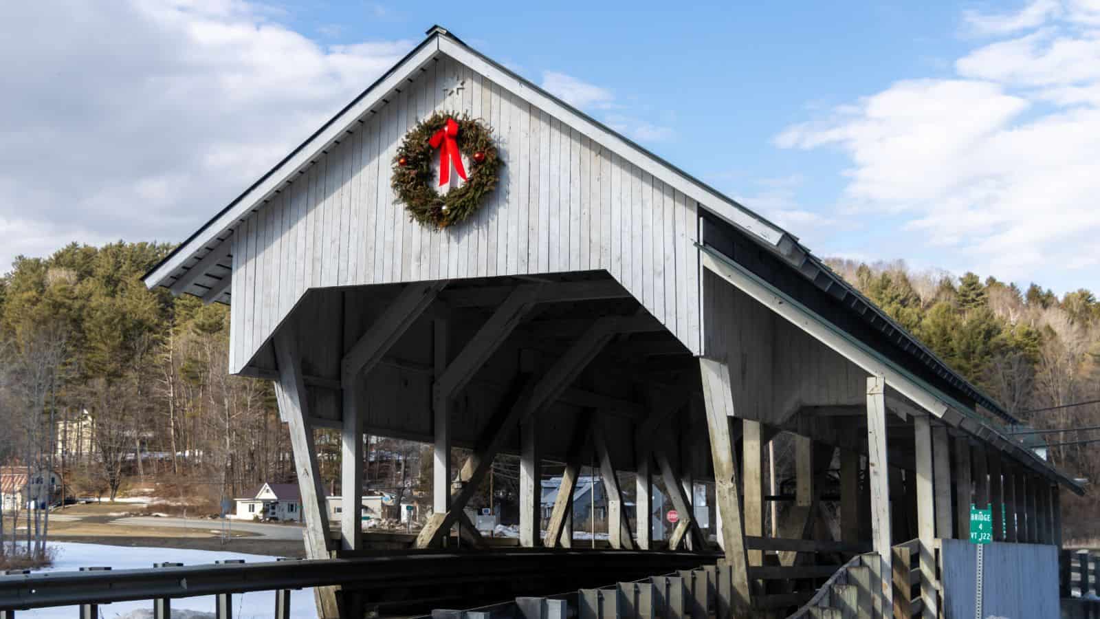 A white wooden covered bridge decorated with a wreath and red bow, set in a rural area with trees and houses in the background.