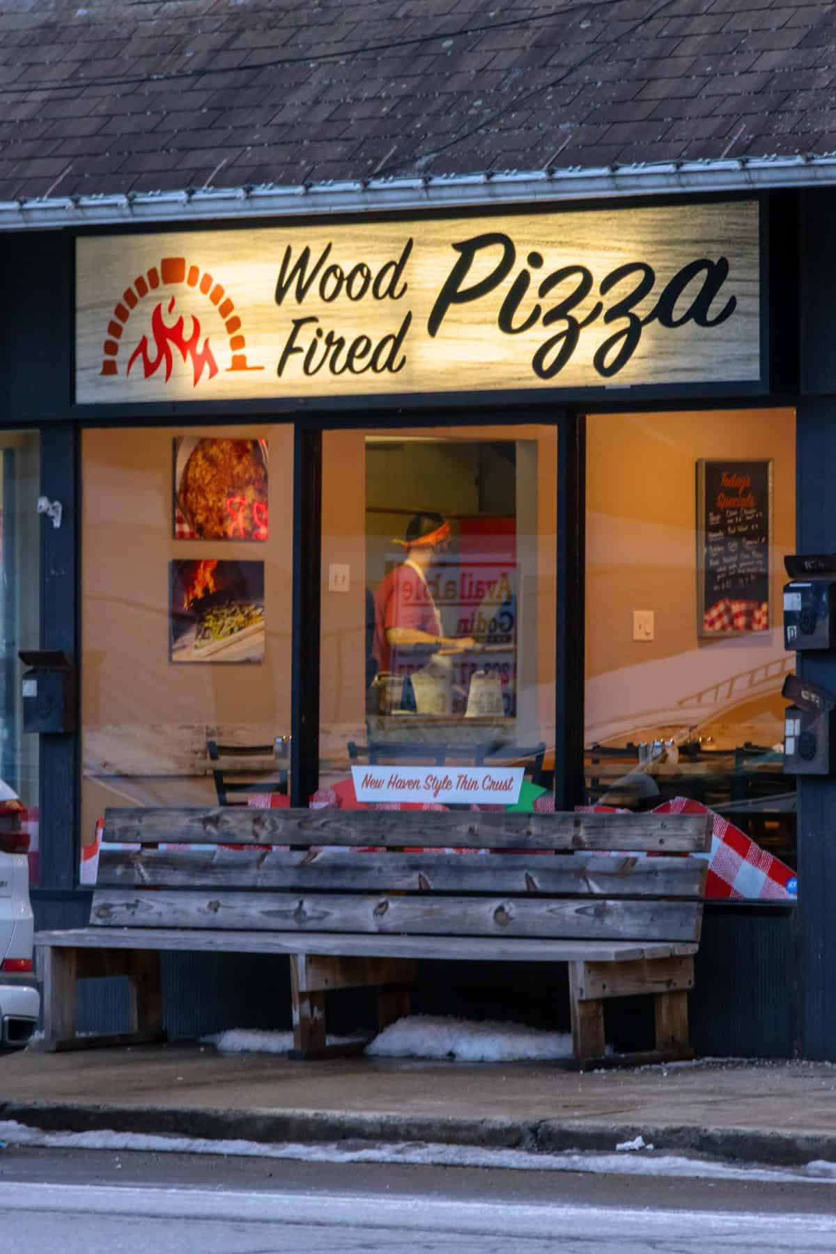 A storefront with a sign reading "Wood Fired Pizza," a window showing a person inside, benches outside, and a small patch of snow on the ground.