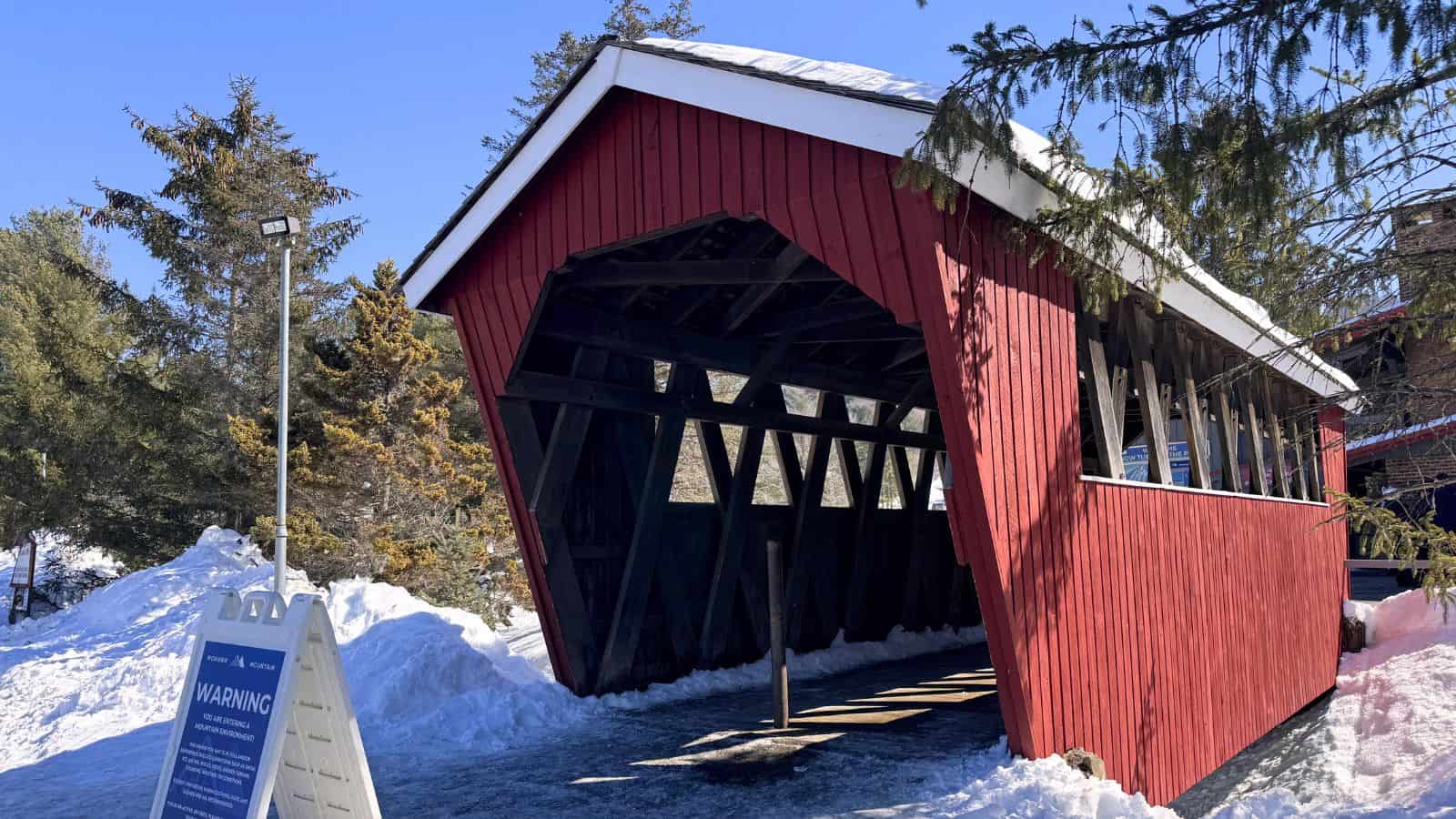 A red covered wooden bridge stands over a snow-covered path, surrounded by evergreen trees and warning signs.