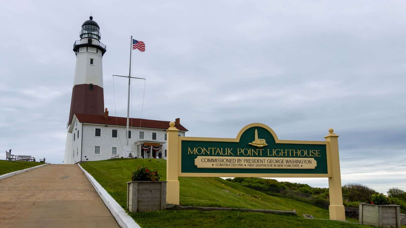Montauk Point Lighthouse stands on a grassy hill under a cloudy sky, with an American flag and a green sign detailing its historical significance in the foreground.