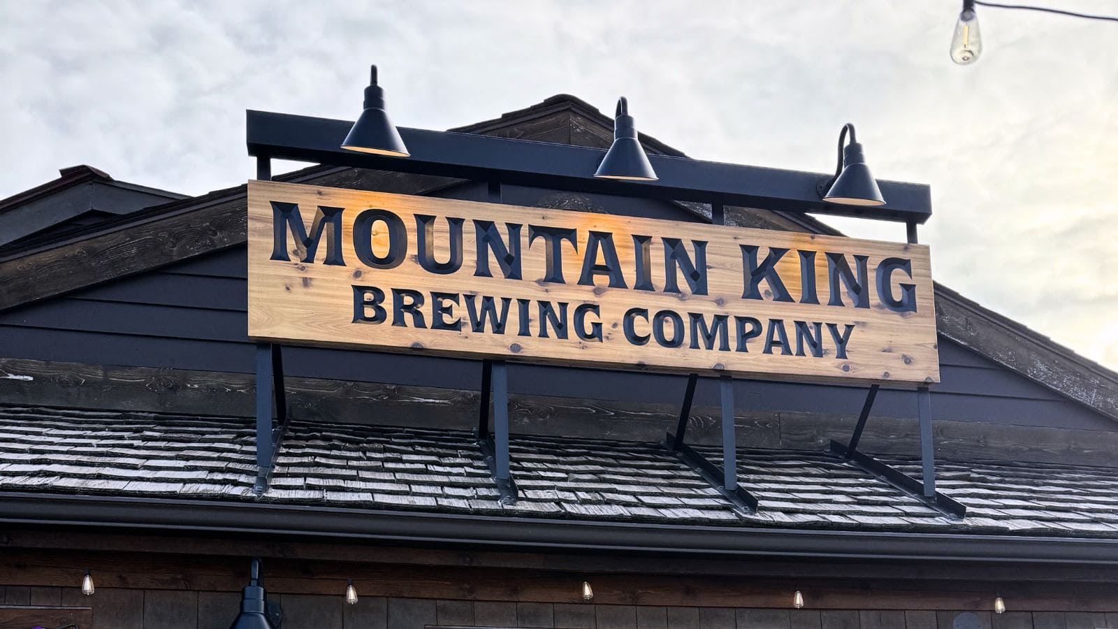 Wooden sign reading "Mountain King Brewing Company" mounted above the entrance of a building with three exterior lights illuminating the sign.