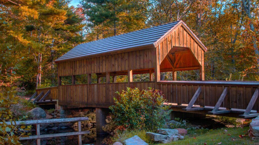 A wooden covered bridge spans a small stream amid trees with autumn foliage.