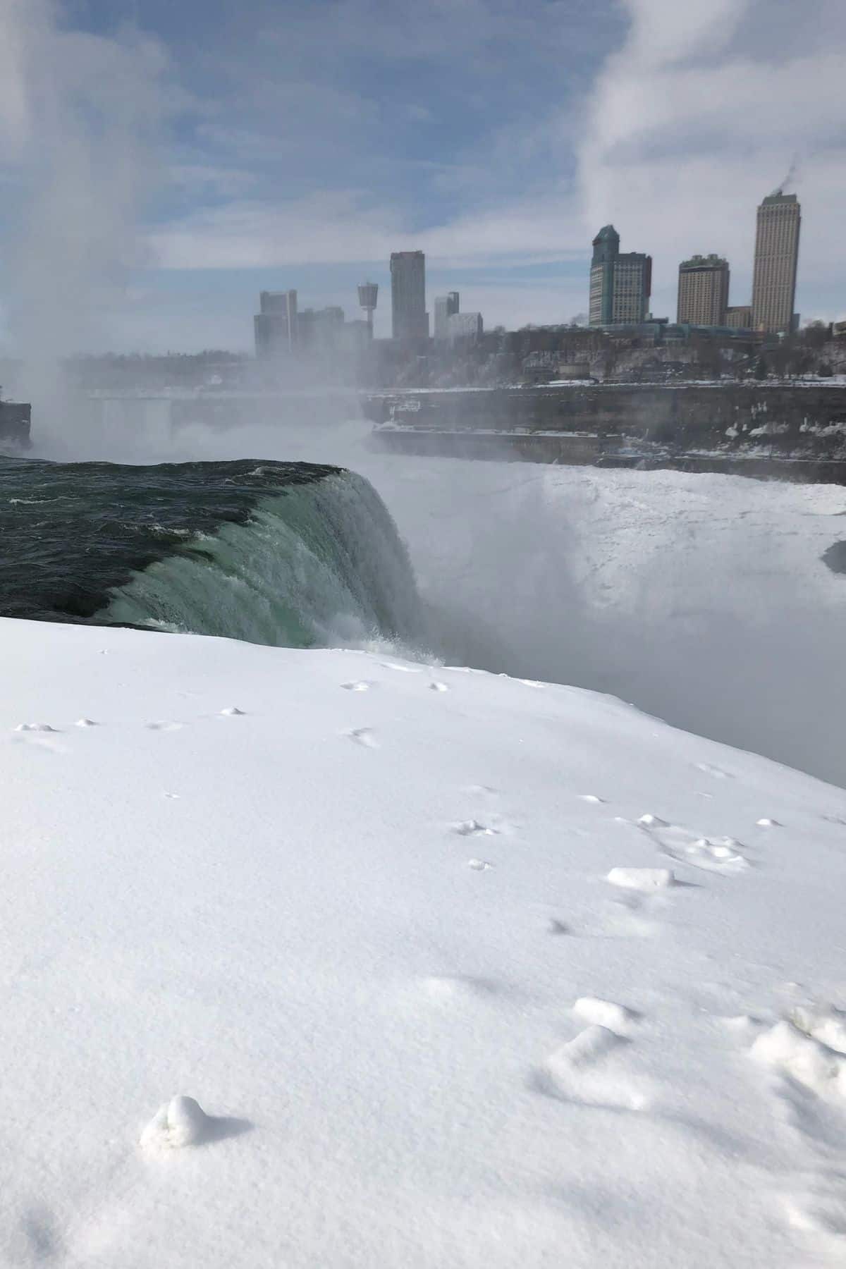 Snow covers the ground near the edge of Niagara Falls, with mist rising and city buildings visible in the background under a partly cloudy sky.
