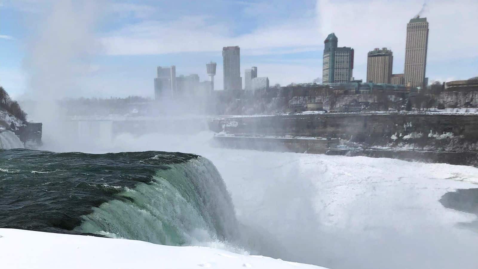 A view of Niagara Falls with water cascading over the edge, mist rising, and city buildings visible in the background under a partly cloudy sky.