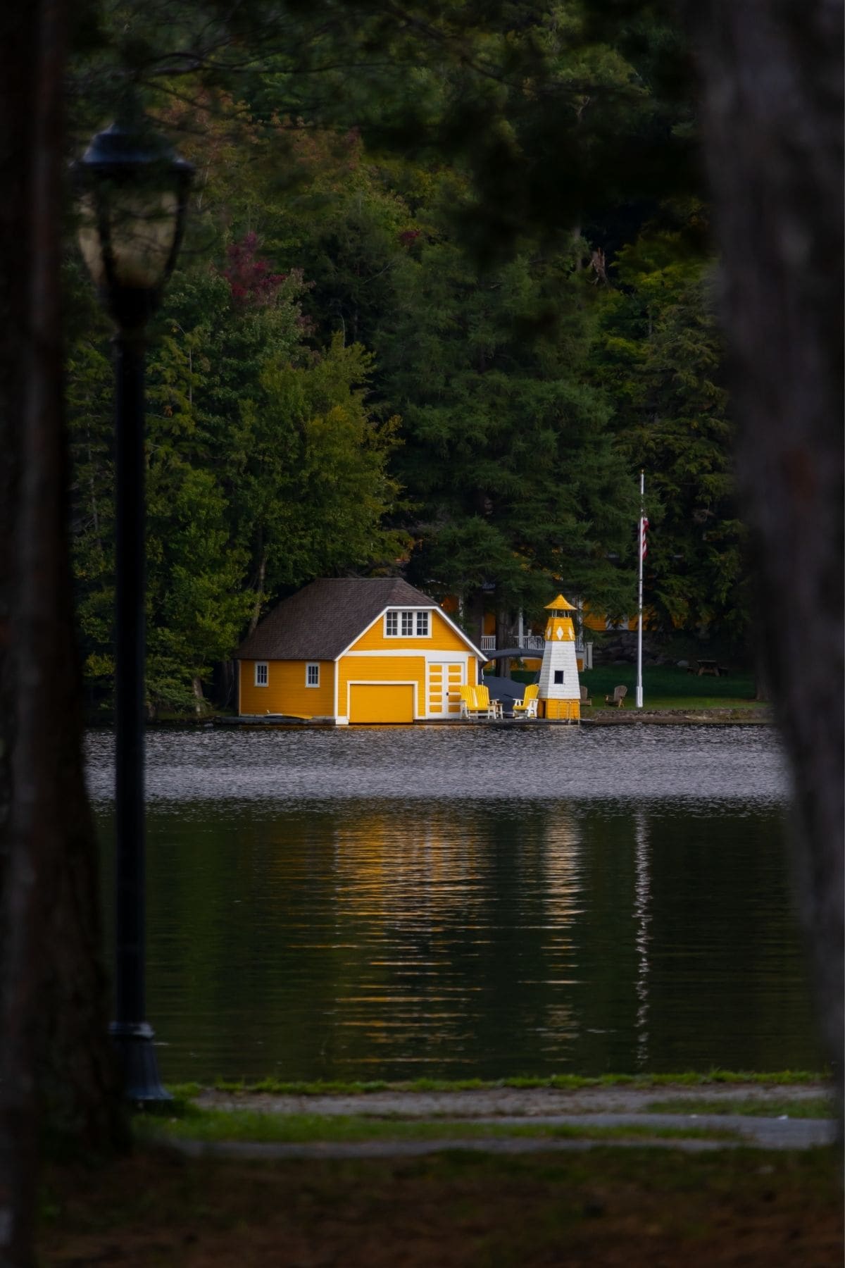 A yellow boathouse and small lighthouse sit on the edge of a calm lake, surrounded by trees, with their reflection visible in the water.