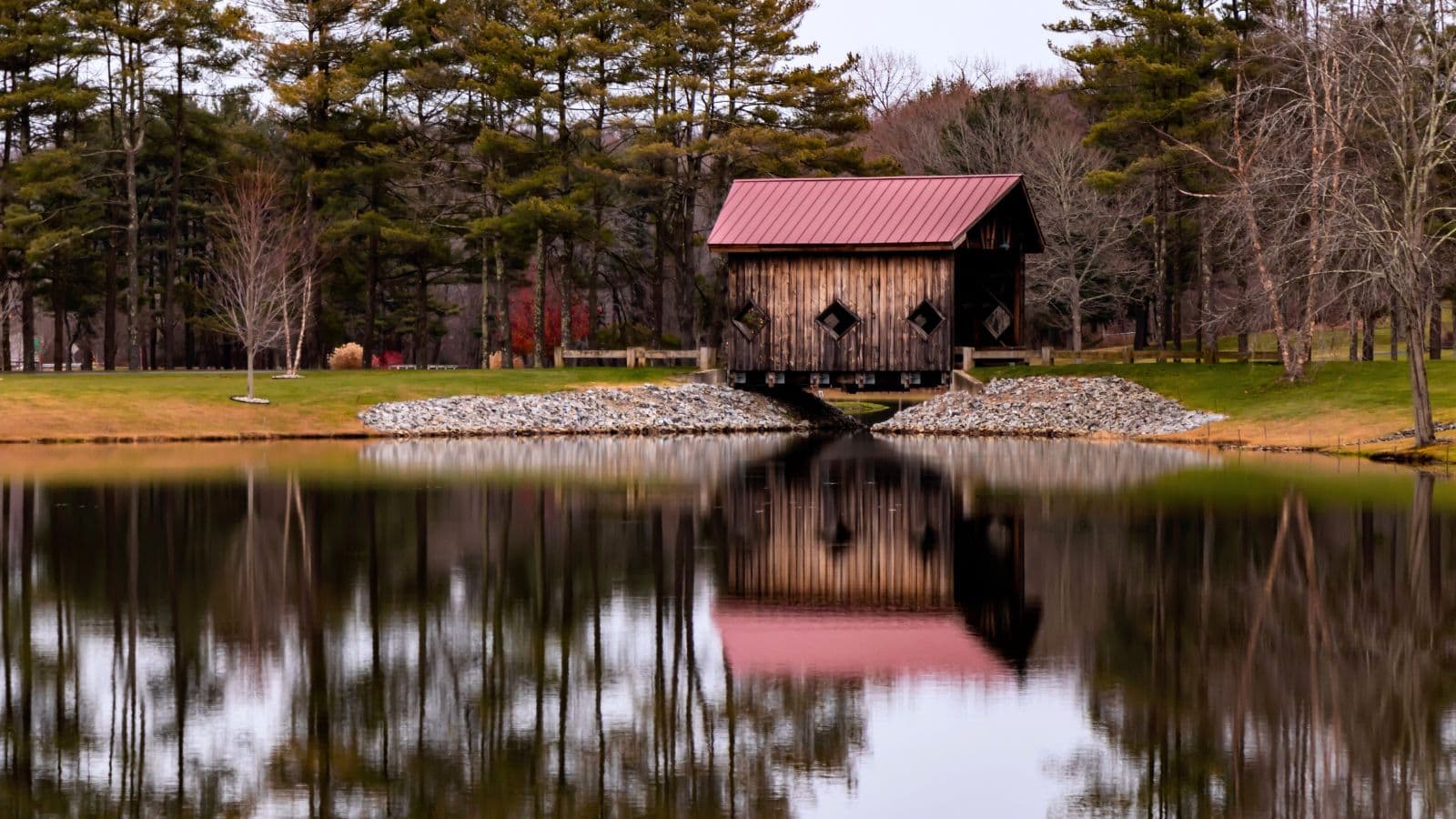 A small wooden building with a red roof stands by a pond, surrounded by trees, with its reflection visible in the water.