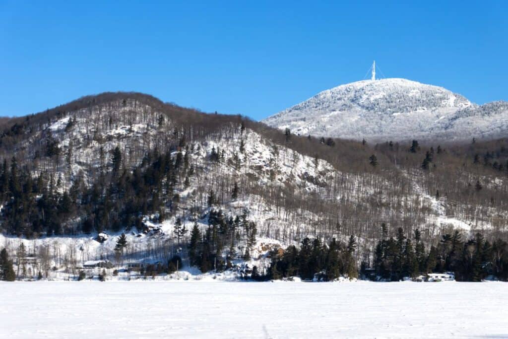Snow-covered hills and mountain with a visible antenna tower on the peak, under a clear blue sky, and a frozen landscape in the foreground.