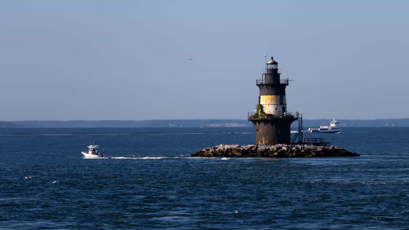 A lighthouse stands on a rocky island in the sea, with boats passing nearby under a clear sky.